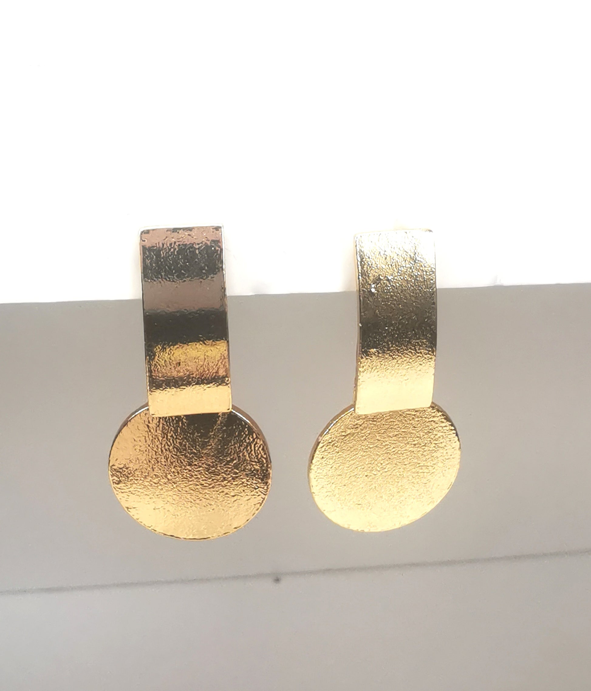 Boucles d'oreilles sur clous en métal doré. Un premier rectangle vertical est fixé au clou. En dessus est suspendu un disque. Le tout est texturé. Présentées cote à cote, accrochées sous un support blanc. Vue de face.