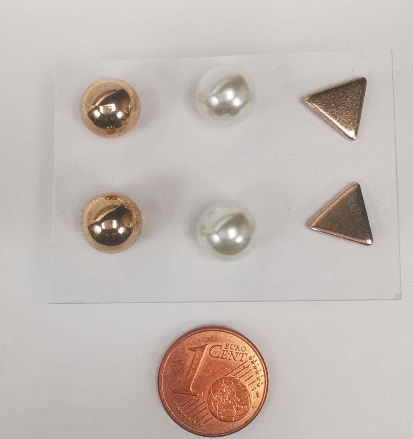 Trios de boucles d'oreilles clous fantaisie, la première est un triangle doré, la deuxième une perle fantaisie et la troisième une sphère dorée. Présentées plantées sur un fond blanc, au dessus d'une pièce de 1 centime, vue de dessus.