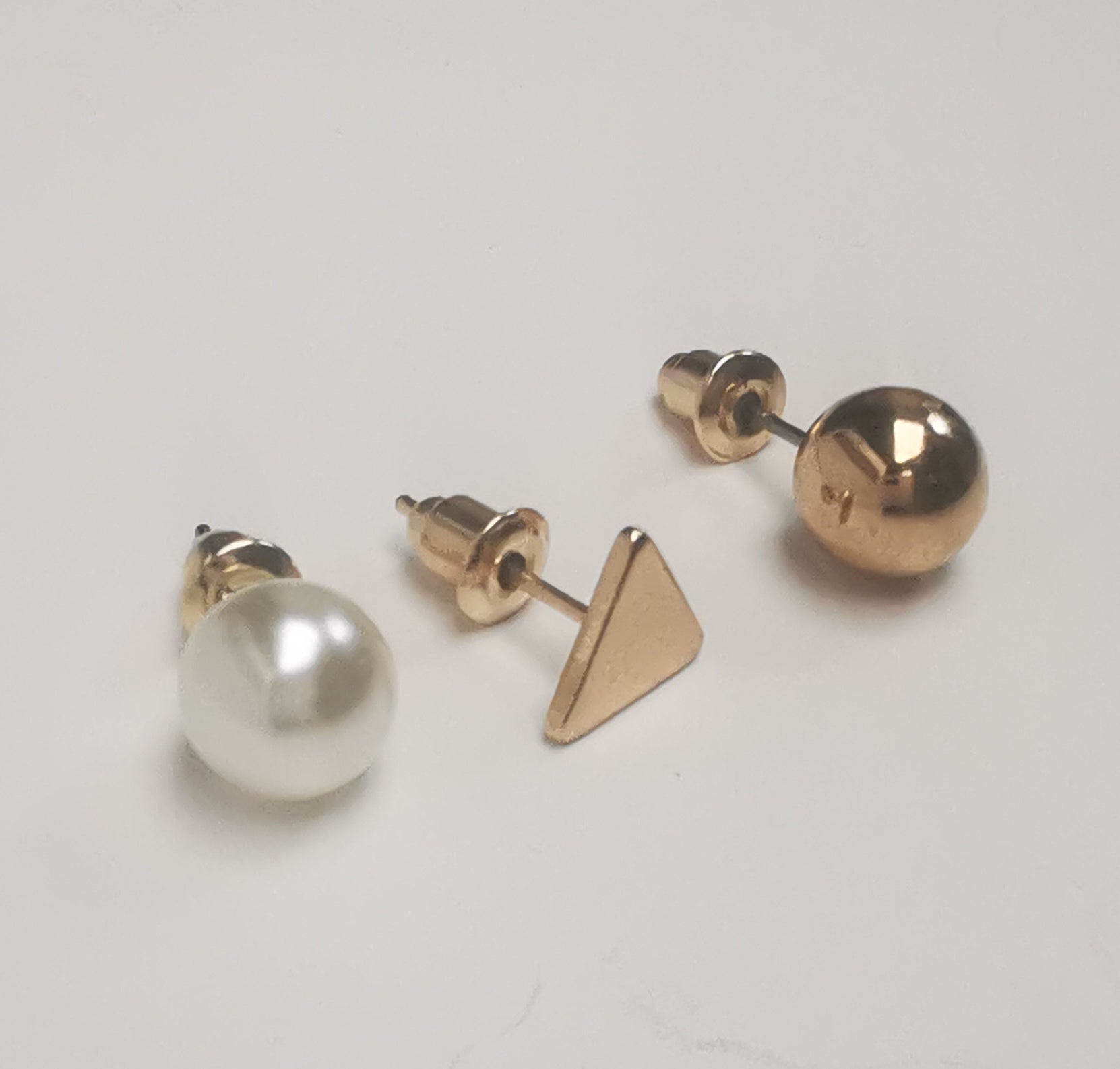 Trios de boucles d'oreilles clous fantaisie, la première est un triangle doré, la deuxième une perle fantaisie et la troisième une sphère dorée. Représentées par une boucle de chaque paire, posées les unes à coté des autres, posées sur un fond blanc, vue de face un peu de gauche.