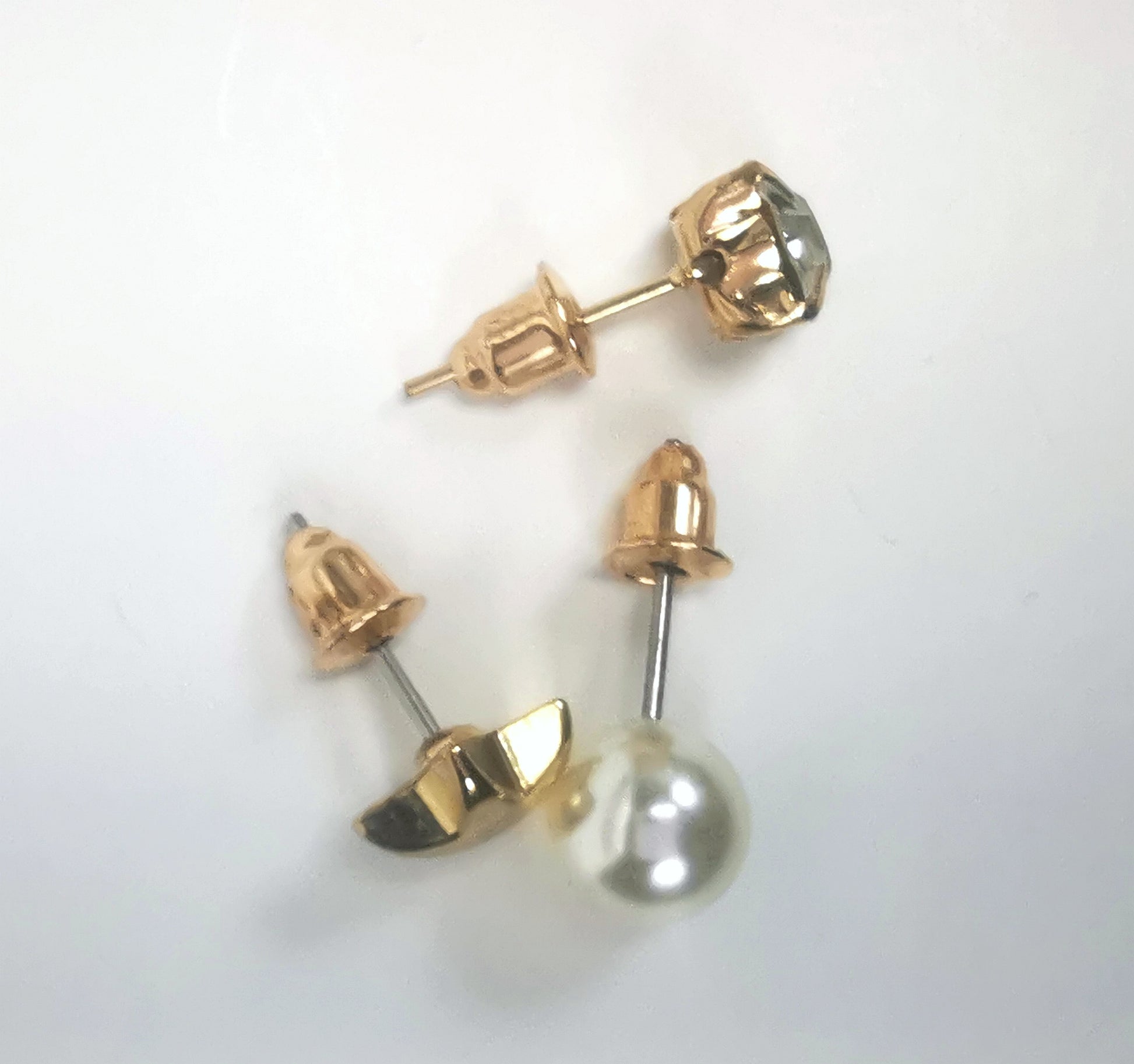 Trios de boucles d'oreilles clous fantaisie dorées, la première est une étoile dorée, la deuxième un faux diamant en strass et la troisième une perles fantaisie blanche, les trois de 7mm. Représentées par une boucle de chaque paire, posées en triangle, posées sur un fond blanc, vue de dessus.