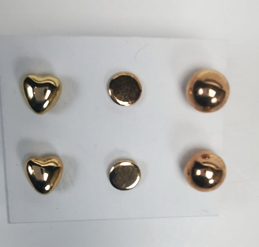 Trois paires de boucles d'oreilles clous dorées, présentées sur fond blanc. Chaque paire est présentée une boucle au dessus de l'autre. La paire de gauche est un coeur, celle du centre un disque plat, celle de droite une sphère . Vue de dessus.