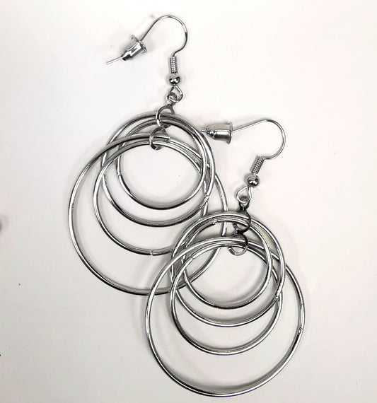 Boucles d'oreilles pendantes argentées de 60mm de long. Composées de quatre anneaux imbriqués les uns dans les autres, Le tout argenté est présenté en paire, posées sur un fond blanc, cote à cote, la boucle de gauche décalée vers le haut. Vue de dessus.