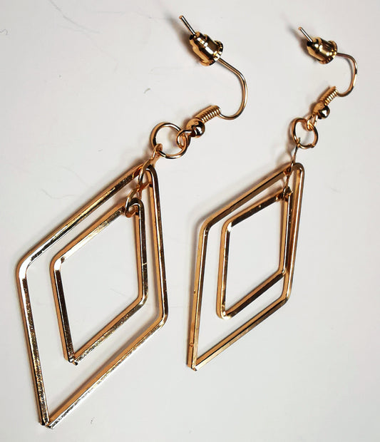 Boucles d'oreilles pendantes dorées de 65mm de long. Composées de 2 losanges imbriqués l'un dans l'autre, Le tout doré est présenté en paire, posées sur un fond blanc, cote à cote, la boucle de droite décalée vers le haut.. Vue de dessus depuis la gauche