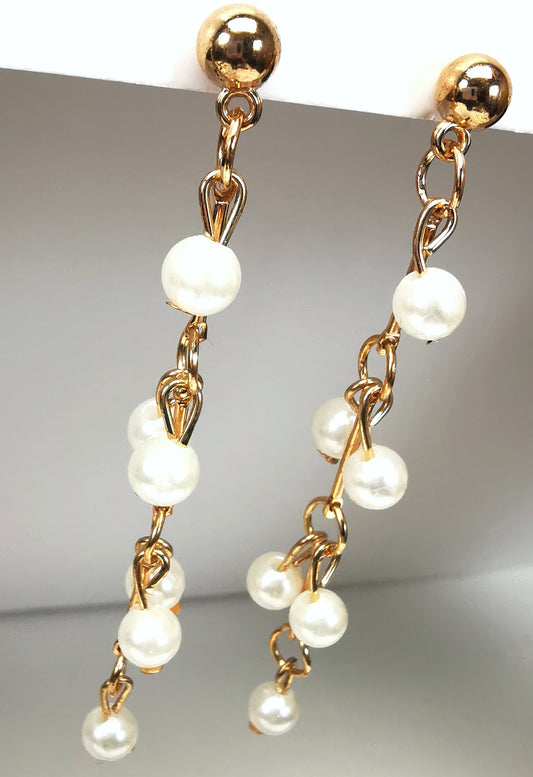 Boucles d'oreilles pendantes dorées sur clous, constituées d'une chaine de 75mm, agrémentée  de perles blanches fantaisie tous les 20mm. Présentées en paire accrochées sous un support, vue légèrement de gauche.