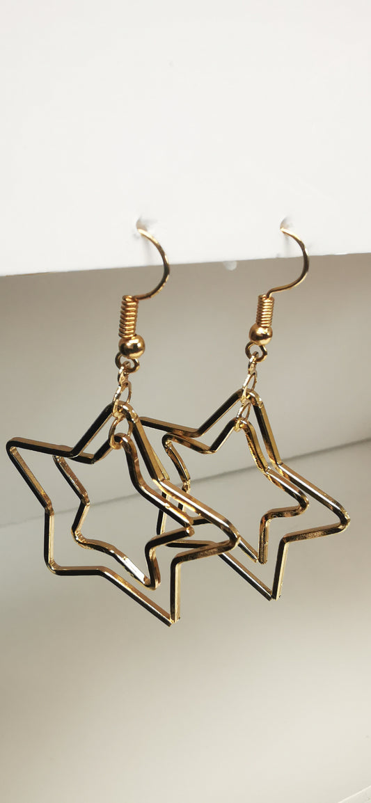 Boucles d'oreilles pendantes dorées de 65mm de long. Composées de 2 étoiles imbriquées l'une dans l'autre, Présenté en paire, suspendues sous un support, cote à cote. Vue de face haut.