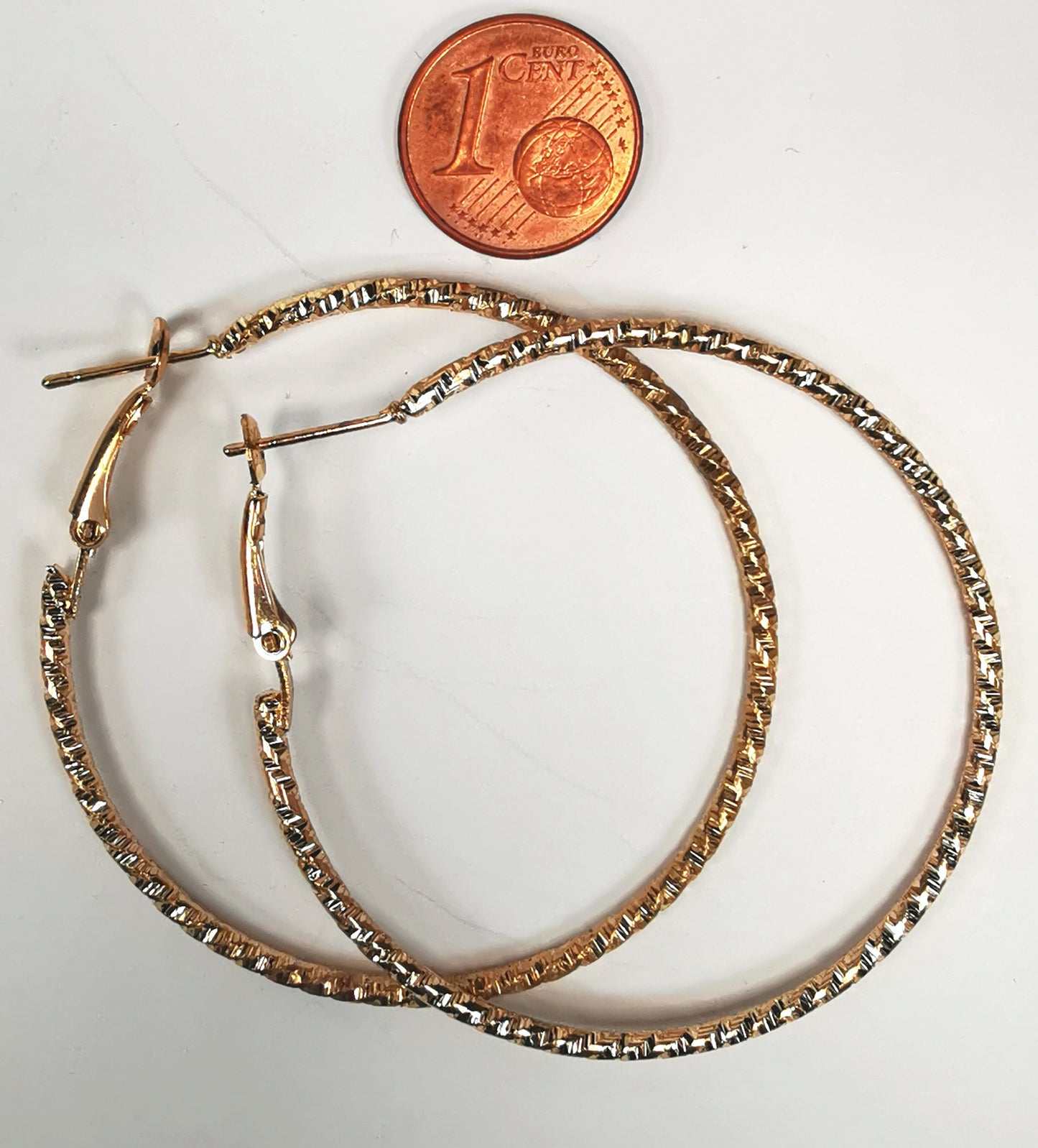 Boucles d'oreilles créoles dorées 50mm de diamètre, fil de 1.5mm, surface texturée. Posées sur fond blanc l'une sur l'autre avec, au dessus, une pièce de 1 centime, vue de dessus.