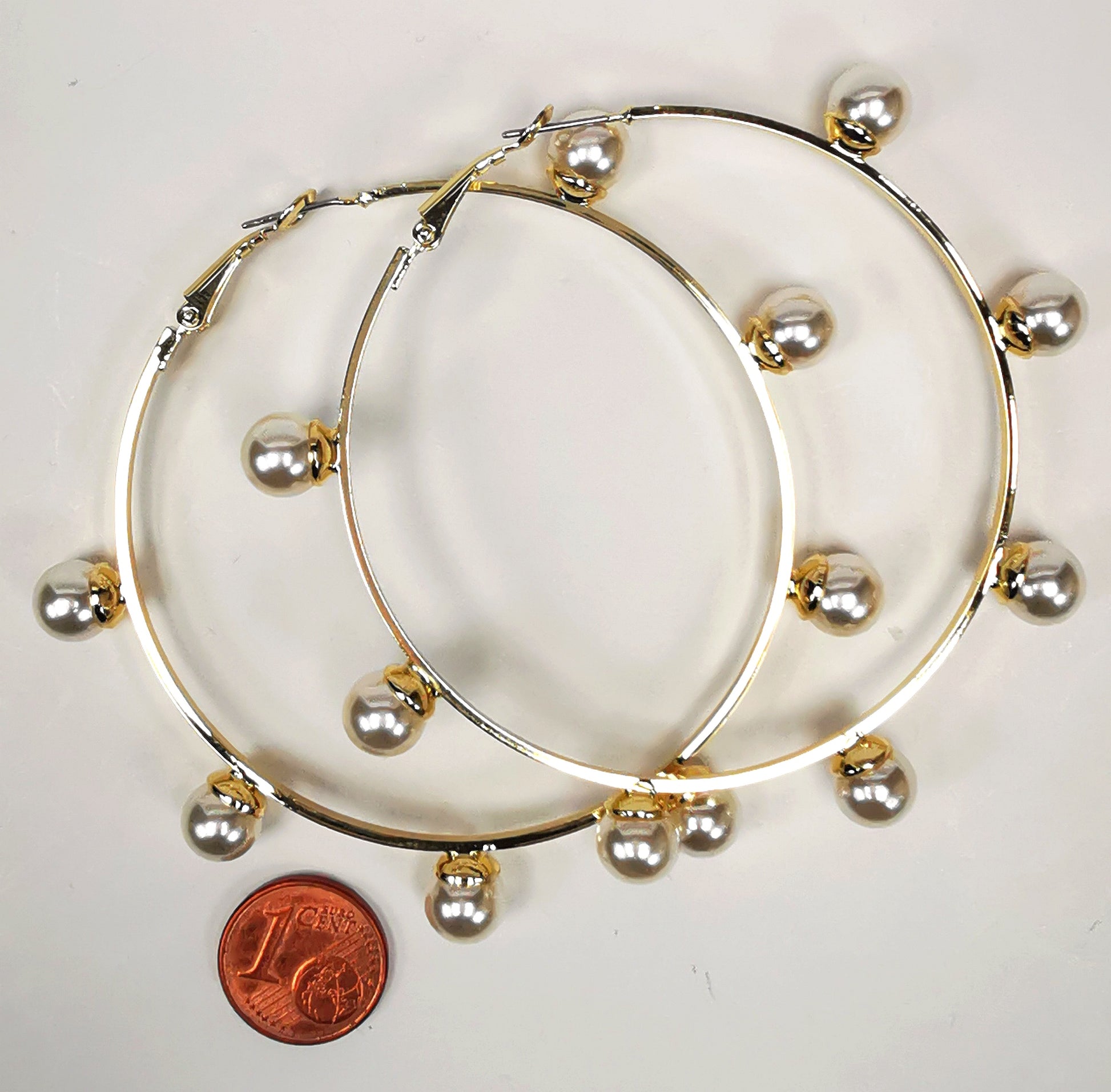 Boucles d'oreilles créoles dorées, diamètre 63mm avec 6 perles fantaisie de 8mm sur l'extérieur. présentées posées sur un fond blanc, l'une sur l'autre, au dessus d'une pièce de 1 centime. Vue de dessus.