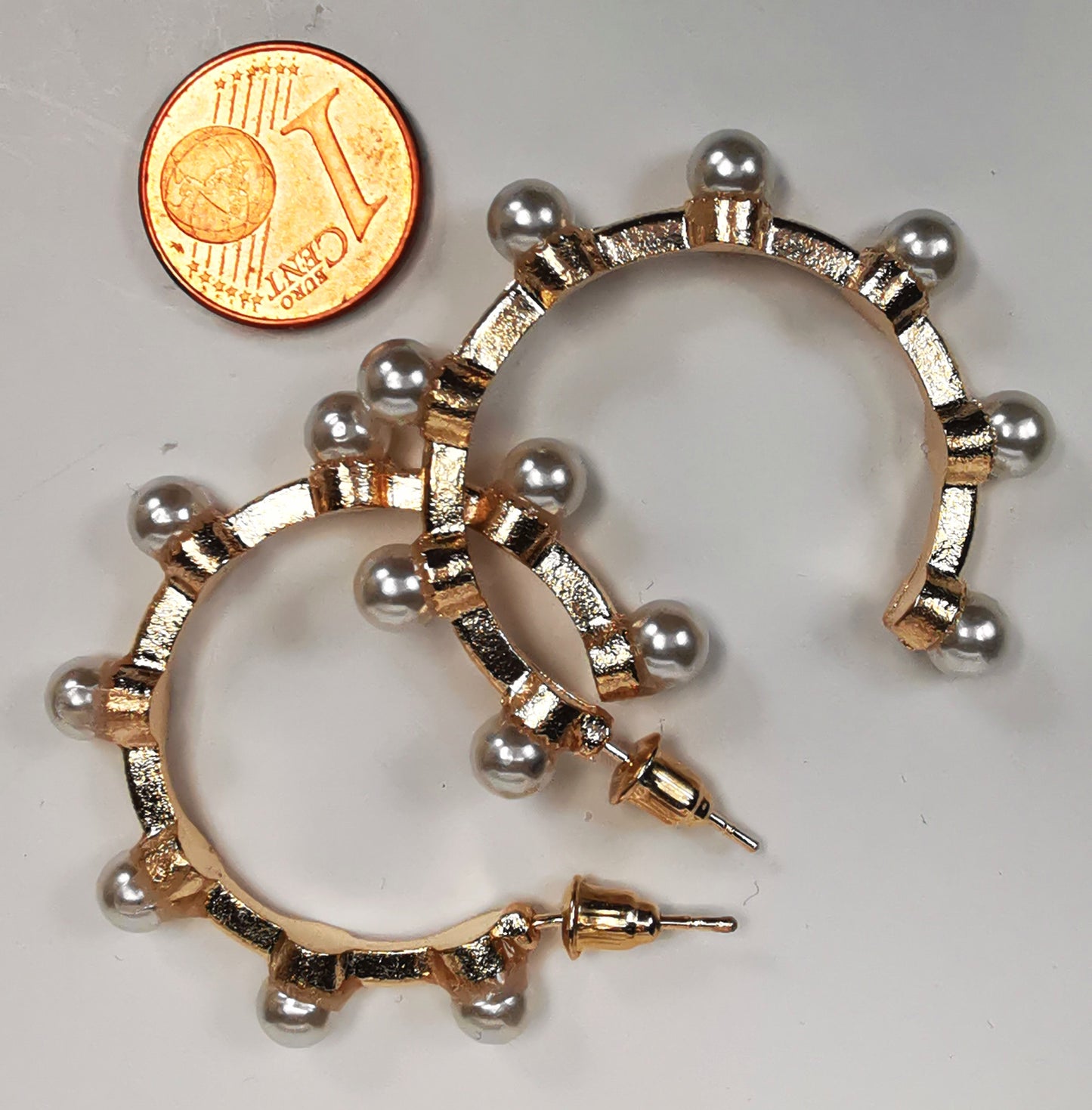 Boucles d'oreilles créoles fantaisie, en alliage doré, 35mm de diamètre, sur clous, avec 8 perles fantaisie sur la périphérie. Présentées l'une à coté de l'autre, posées sur fond blanc à coté d'une pièce de 1 centime. Vue de dessus.