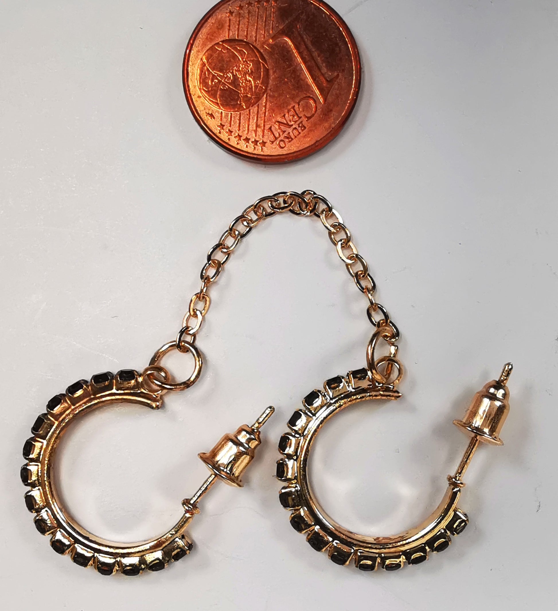 Boucles d'oreilles créoles fantaisie 20mm trois quarts de tour sur clous, posées l'une à coté de l'autre dans le même sens, avec une pièce de 1centime au dessus. Chacune est composée d'un anneau doré incrusté se strass noirs sur toute la périphérie. Les deux boucles sont liées par une chaîne dorée d'environ 35mm, se portent sur la même oreille