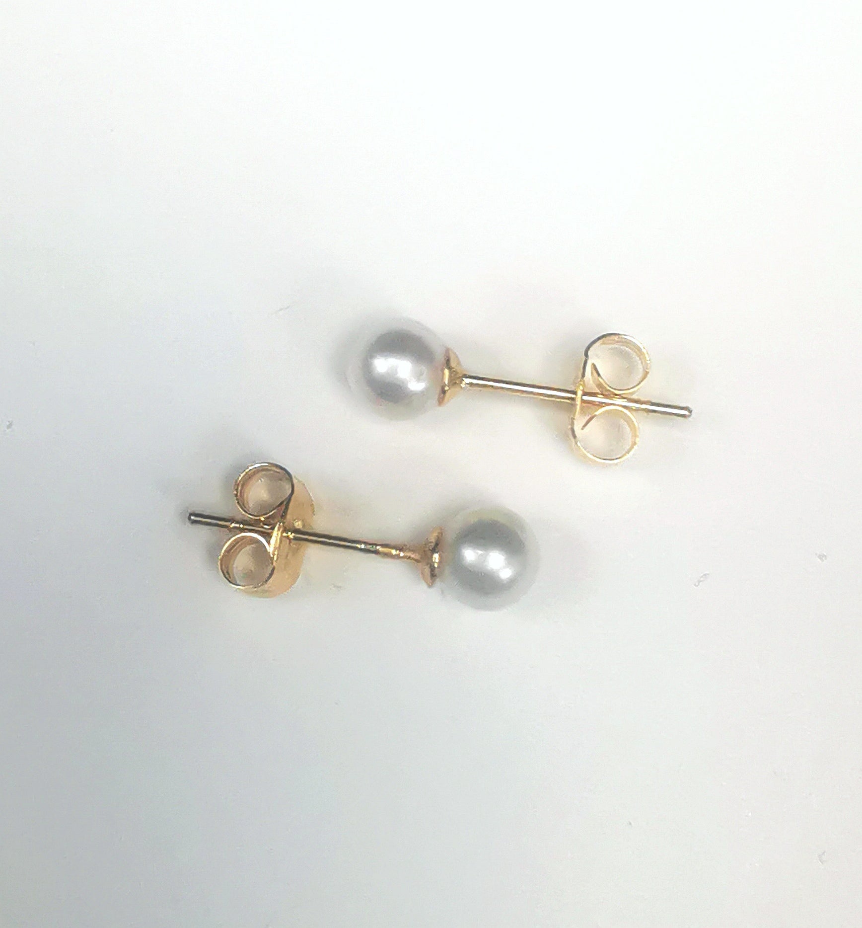 Boucles d'oreilles perles de culture blanches rondes 5mm montées sur clous plaqués or. Présentées horizontalement, celle de dessus perle à gauche, celle de dessous perle à droite, sur fond blanc. Vue de dessus.