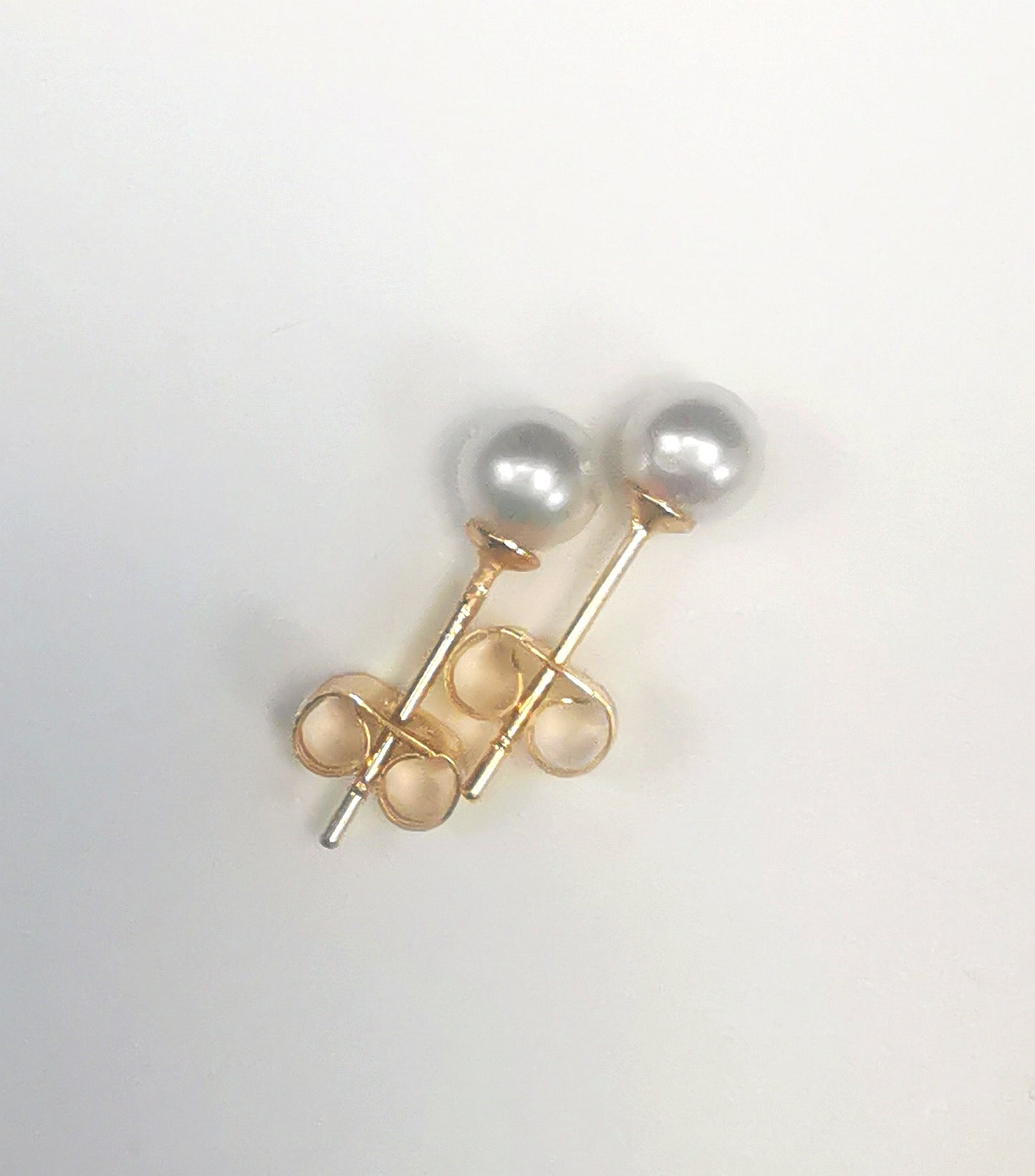 Boucles d'oreilles perles de culture blanches rondes 5mm montées sur clous plaqués or. Présentées cote à cote, perles en haut, les deux décalées vers la droite, sur fond blanc. Vue de dessus.