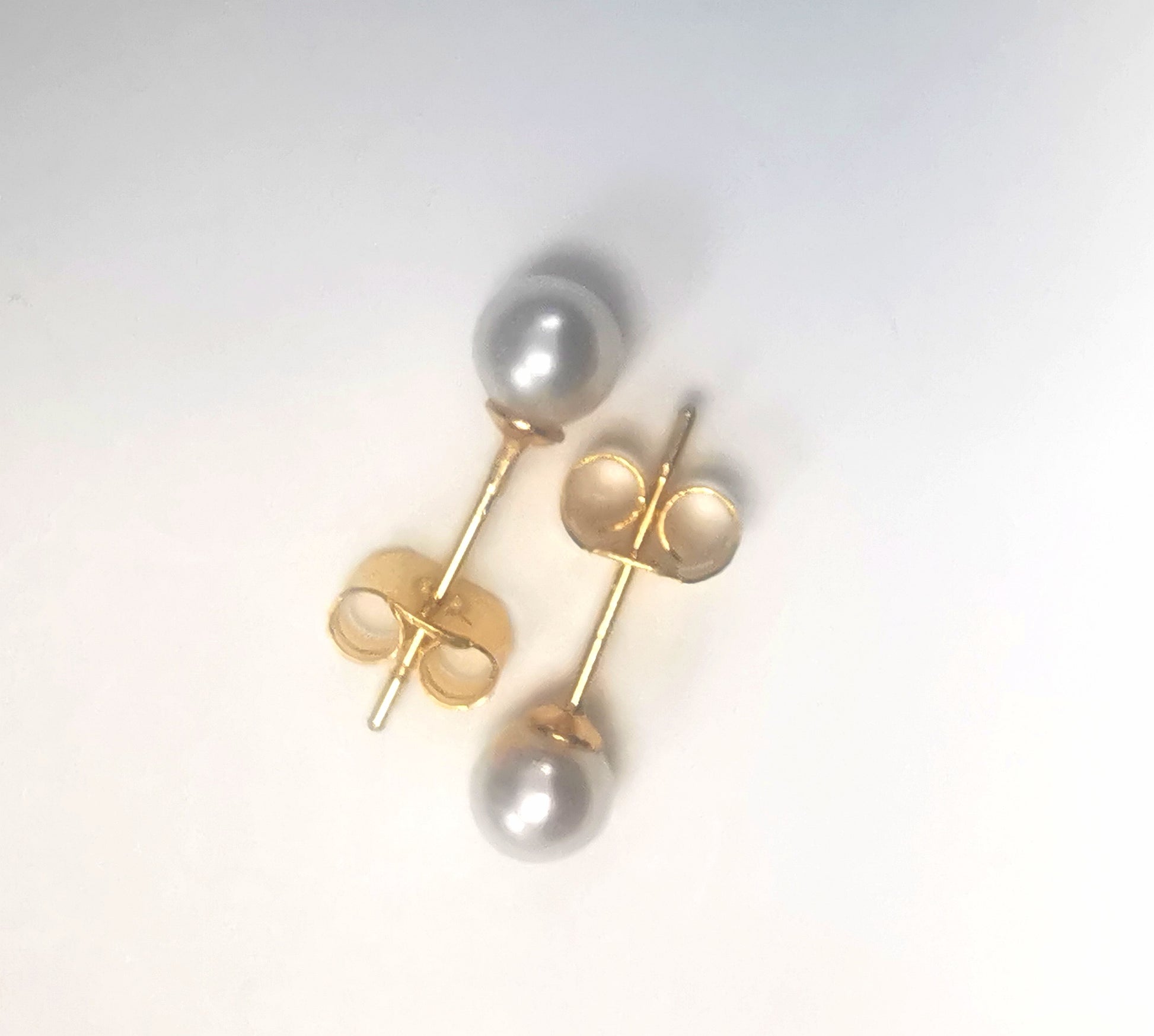 Boucles d'oreilles perles de culture blanches rondes 5mm montées sur clous plaqués or. Présentées cote à cote, têtes bèches, boucles de gauche perle en haut, celle de droite, perle en bas, sur fond blanc. Vue de dessus.