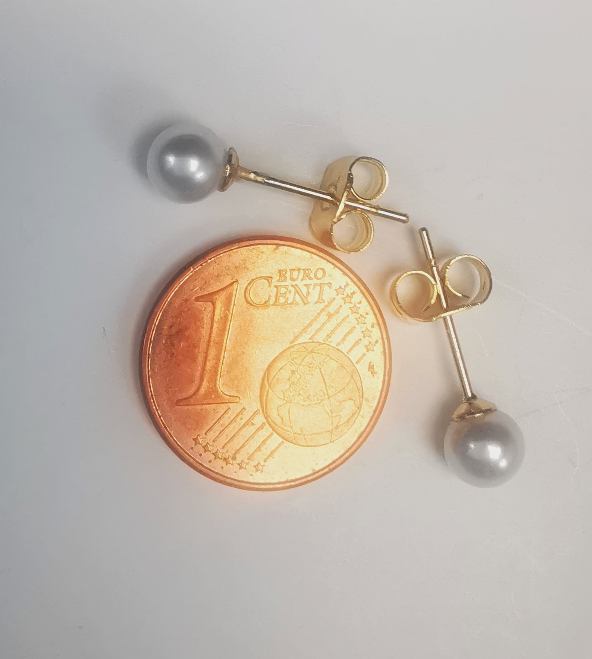 Boucles d'oreilles perles de culture blanches rondes 5mm montées sur clous plaqués or. Présentées perpendiculairement, celle du haut horizontale perle à gauche, celle du bas verticale perle en bas, au dessus d'une pièce de 1 centime, sur fond blanc. Vue de dessus.