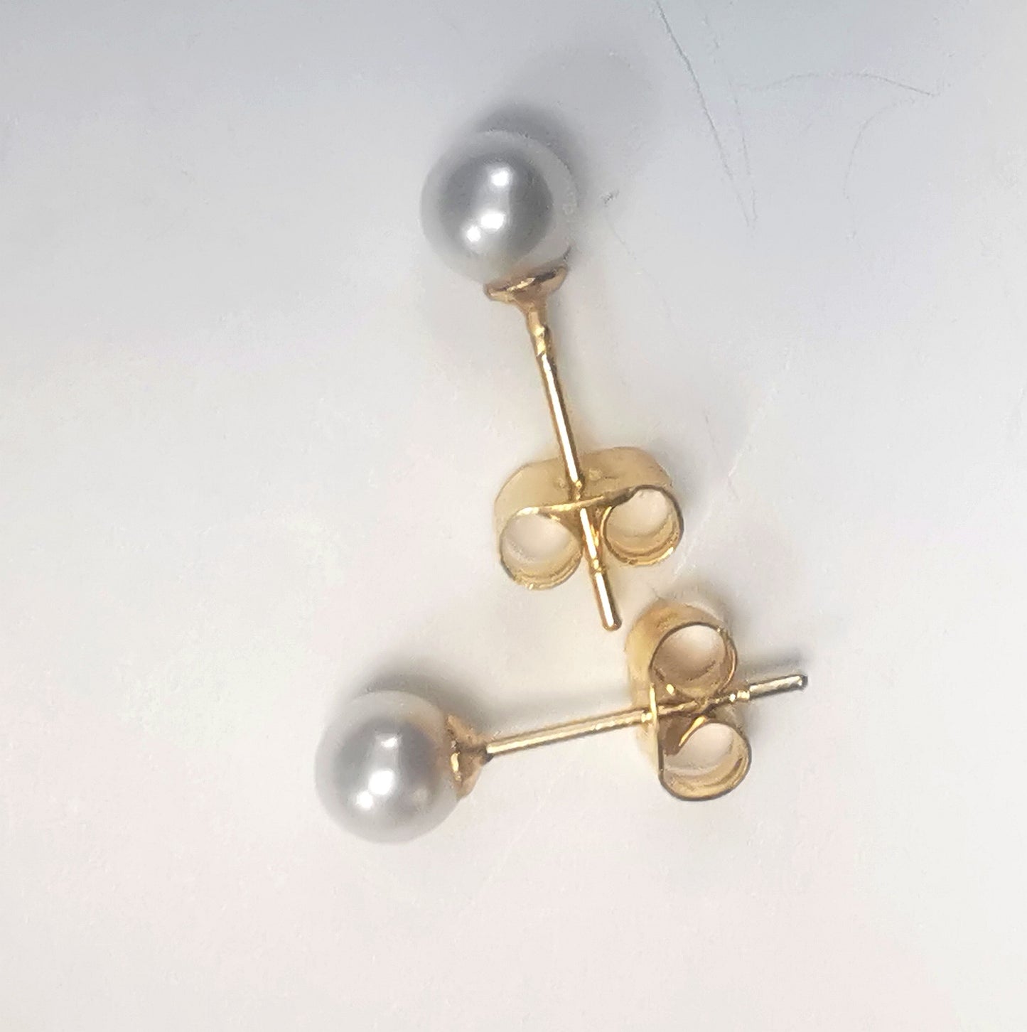 Boucles d'oreilles perles de culture blanches rondes 5mm montées sur clous plaqués or. Présentées perpendiculairement, celle du bas horizontale perle à gauche, celle du haut verticale perle en haut, sur fond blanc. Vue de dessus.