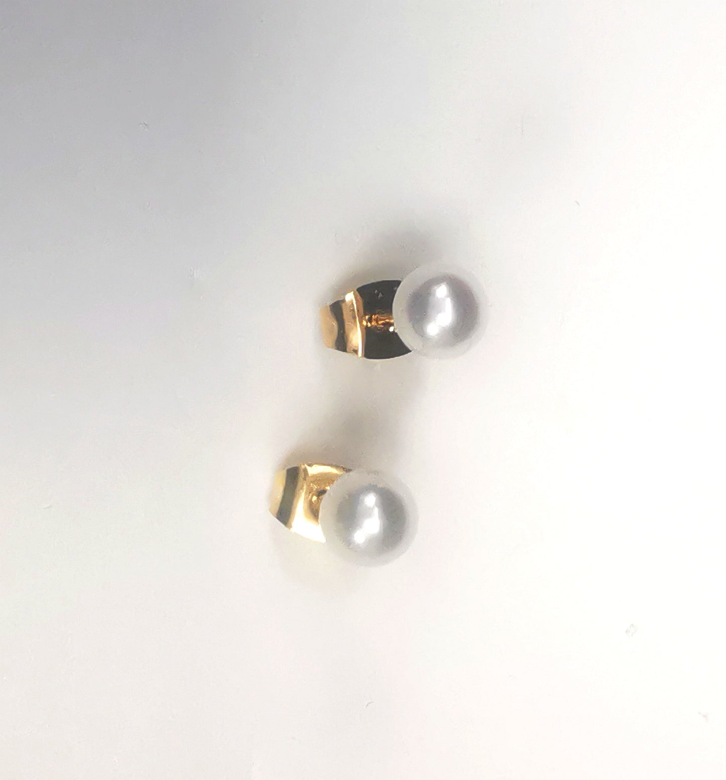 Boucles d'oreilles perles de culture blanches rondes 5mm montées sur clous plaqués or. Présentées platées sur un support blanc, l'une au dessus de l'autre. Vue de dessus.