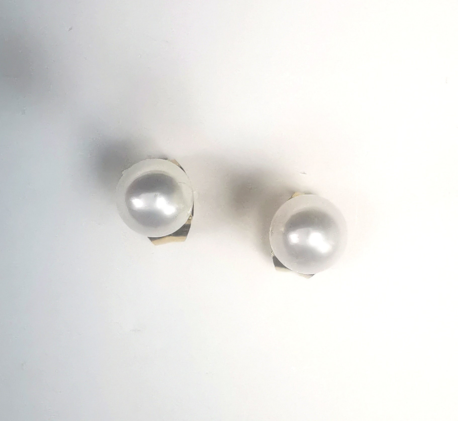 Boucles d'oreilles perles de culture blanches rondes 5mm montées sur clous plaqués or. Présentées platées sur un support blanc, l'une a coté de l'autre. Vue de dessus.