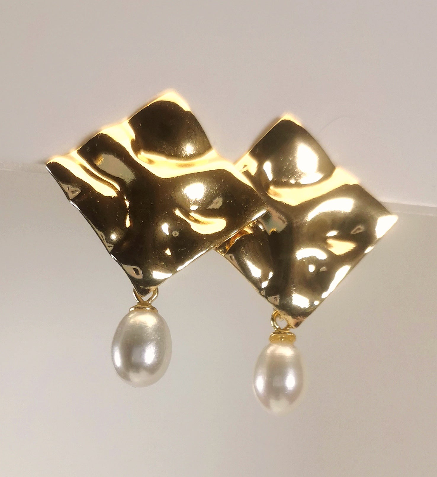 Boucles d'oreilles composées d'un carré martelé de 18mm, plaqué or, sur clou, sous lequel est suspendue une perle blanche , ovale 7-9mm. Présentées posées sur un fond blanc, cote à cote, la boucle de gauche chevauchant celle de droite. Vue  en gros plan de dessus