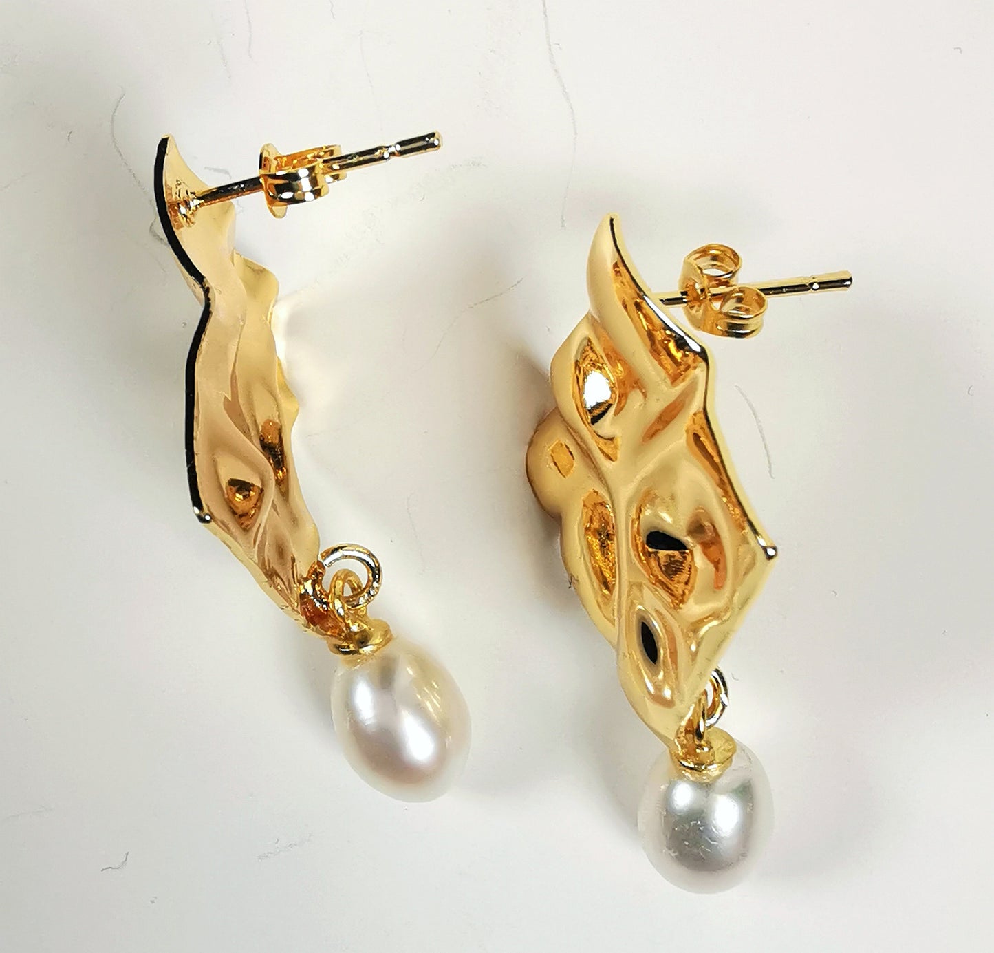 Boucles d'oreilles composées d'un carré martelé de 18mm, plaqué or, sur clou, sous lequel est suspendue une perle blanche , ovale 7-9mm. Présentées posées sur un fond blanc, cote à cote, sur chant Vue de dessus