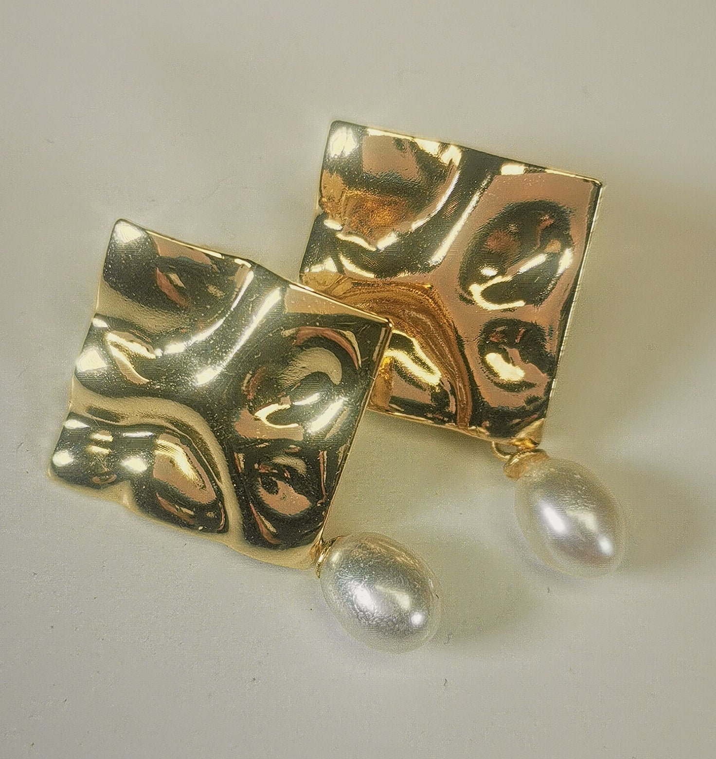Boucles d'oreilles composées d'un carré martelé de 18mm, plaqué or, sur clou, sous lequel est suspendue une perle blanche , ovale 7-9mm. Présentées posées sur un fond blanc, cote à cote, inclinées sur la droite, la boucle de gauche chevauchant celle de droite. Vue en gros plan,de dessus