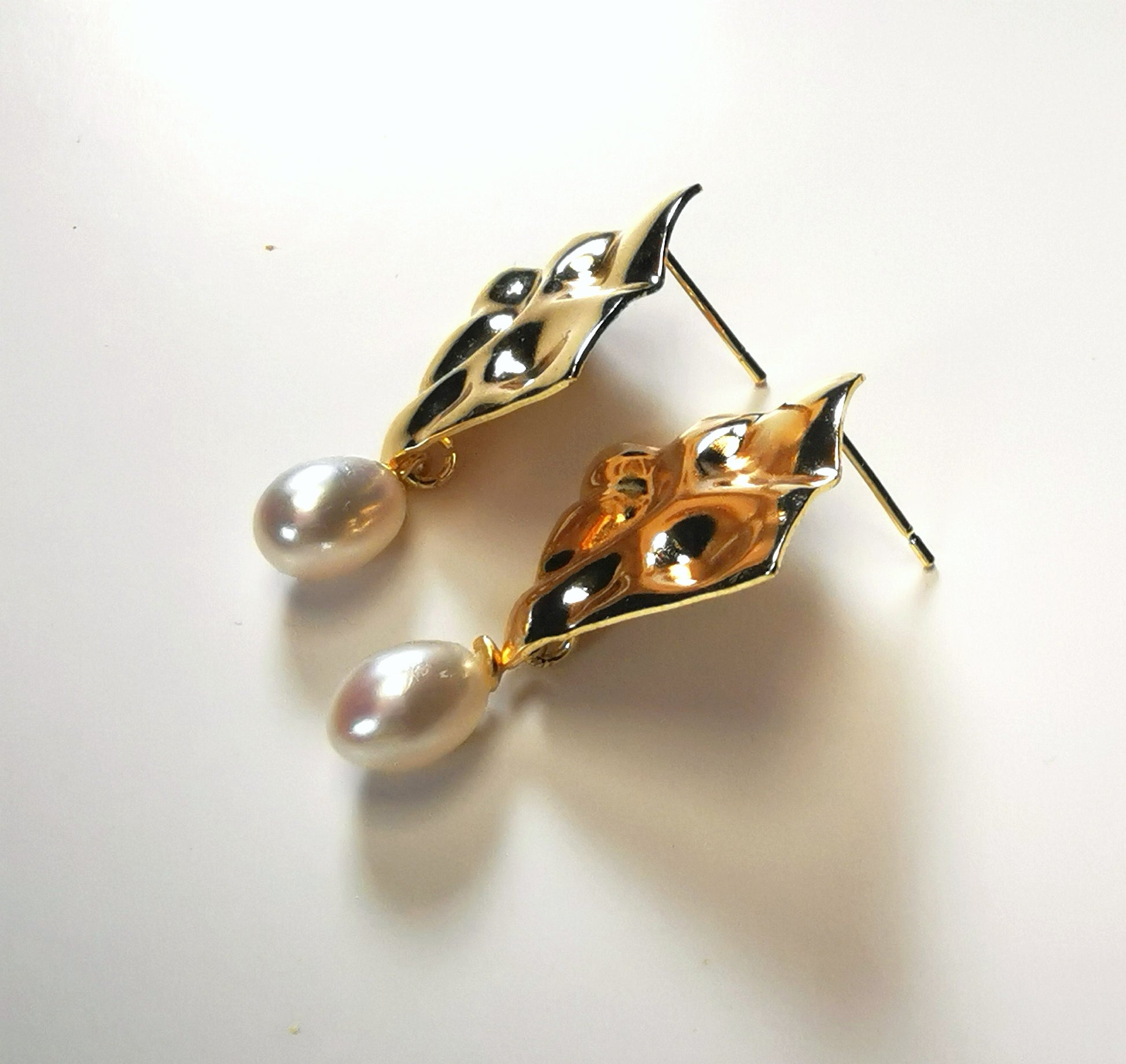 Boucles d'oreilles composées d'un carré martelé de 18mm, plaqué or, sur clou, sous lequel est suspendue une perle blanche , ovale 7-9mm. Présentées posées sur un fond blanc, cote à cote, posées sur le flanc gauche des carrés. Vue de la gauche