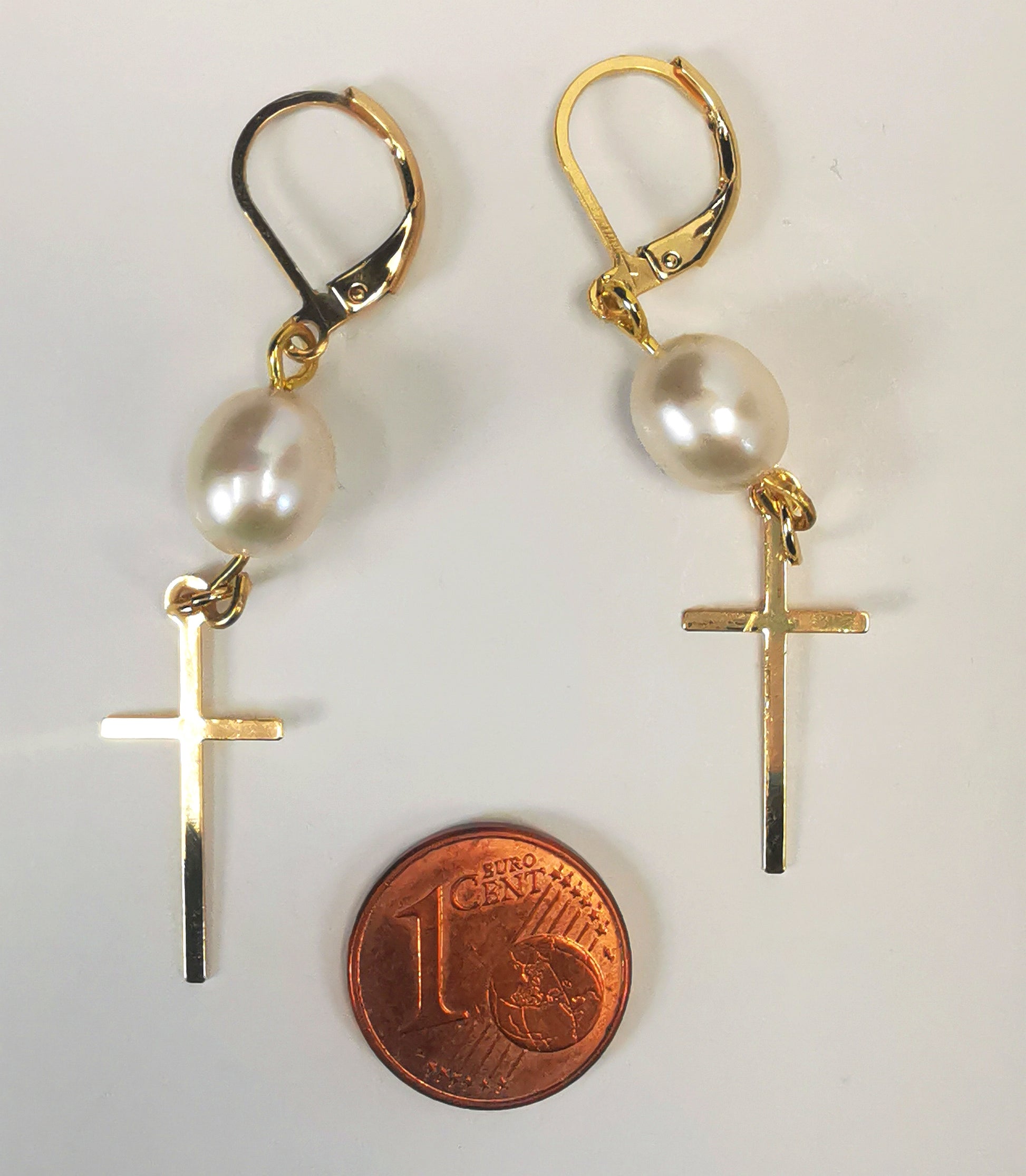 Boucles d'oreilles perles de culture ovale 7-9mm blanche s, accrochées sous dormeuses plaquées or. Sous la perle est suspendue une croix plaquée or de 22x10mm. Présentées posées cote à cote, au dessus d'une pièce de 1 centime, sur un fond blanc. Vue de dessus.