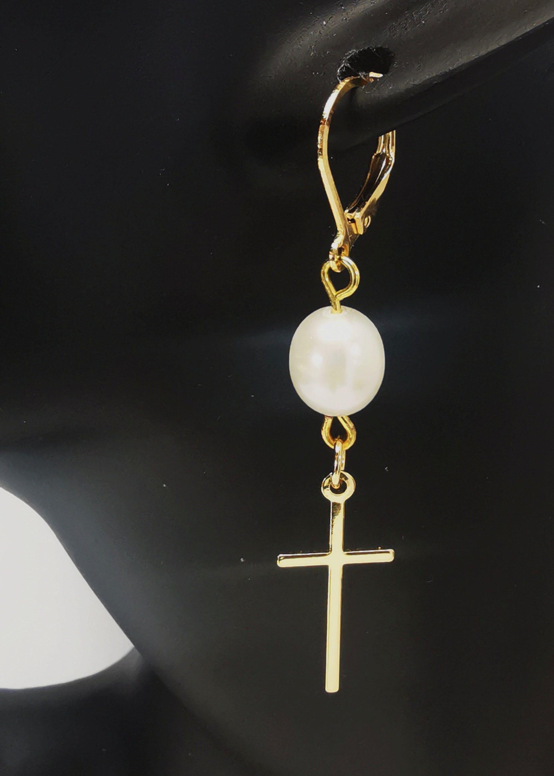 Une boucle d'oreilles perle de culture ovale 7-9mm blanche , accrochée sous dormeuse plaquée or. Sous la perle est suspendue une croix plaquée or de 22x10mm. Présentée accrochée à l'oreille d'un mannequin buste. Vue de coté en très gros plan.