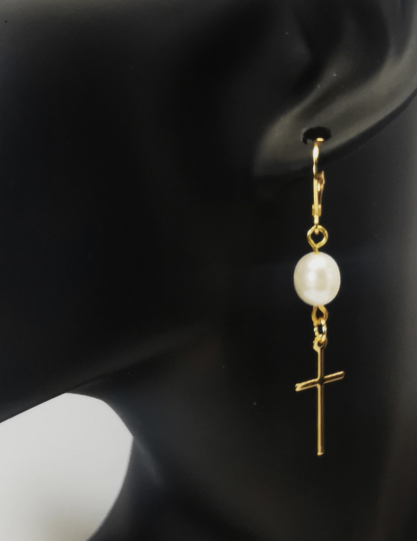 Une boucle d'oreilles perle de culture ovale 7-9mm blanche , accrochée sous dormeuse plaquée or. Sous la perle est suspendue une croix plaquée or de 22x10mm. Présentée accrochée à l'oreille d'un mannequin buste. Vue de trois quart face.