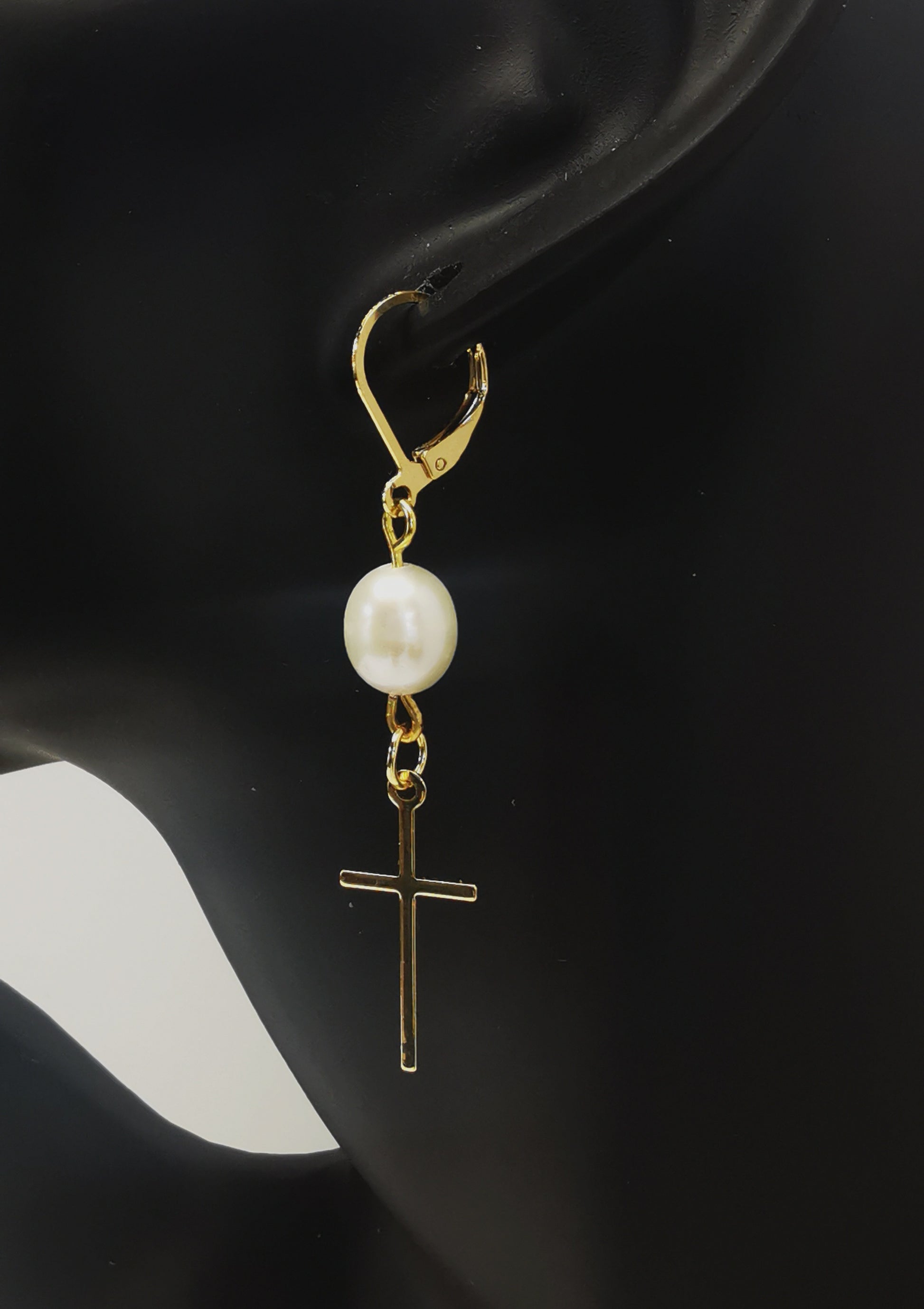 Une boucle d'oreilles perle de culture ovale 7-9mm blanche , accrochée sous dormeuse plaquée or. Sous la perle est suspendue une croix plaquée or de 22x10mm. Présentée accrochée à l'oreille d'un mannequin buste. Vue de coté.