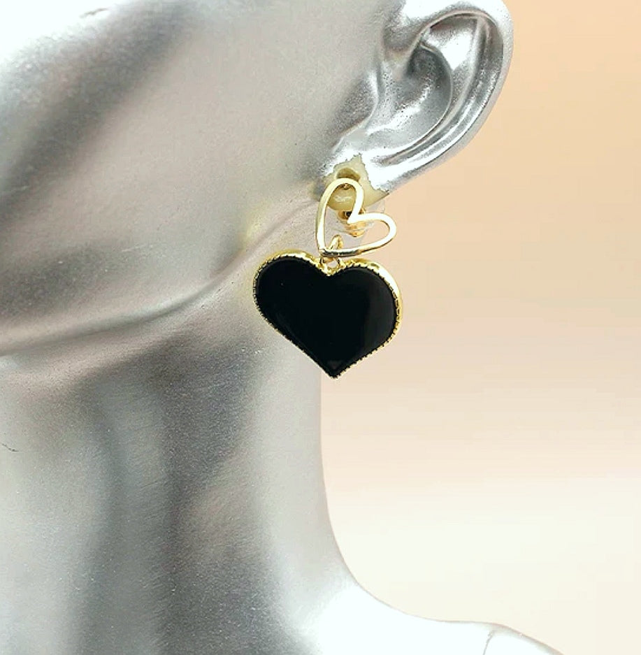Une boucle d'oreille pendante à loreille d'un buste gris, c'est un petit coeur doré sur clou sur lequel pend un coeur noir sur entourage et fond doré